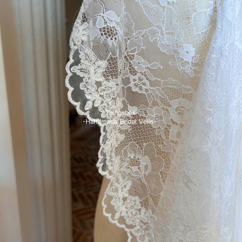 Może przedstawiać: Zbliżenie białego, koronkowego welonu ślubnego. Delikatna koronka ma kwiatowe wzory i falisty brzeg. Widoczny jest tekst "thingsbox - Handmade Bridal Veils".