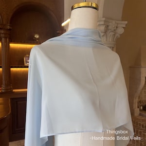 Może przedstawiać: Jasnoniebieski welon ślubny narzucony na manekina. Przezroczysty materiał jest lekki i zwiewny, o miękkim, delikatnym wyglądzie. Tekst "Thingsbox - Handmade Bridal Veils-" jest widoczny w prawym dolnym rogu.