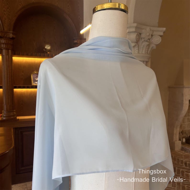 Może przedstawiać: Jasnoniebieski welon ślubny narzucony na manekina. Przezroczysty materiał jest lekki i zwiewny, o miękkim, delikatnym wyglądzie. Tekst "Thingsbox - Handmade Bridal Veils-" jest widoczny w prawym dolnym rogu.