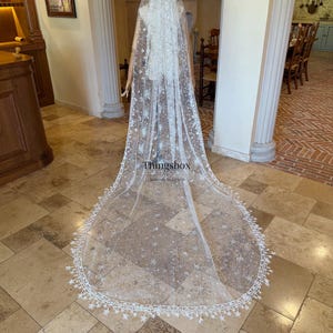 May include: A long, white, sheer bridal veil featuring intricate embroidery of stars, moons, and celestial patterns. The edges are finished with a delicate lace trim shaped like small stars. The text Thingsbox is printed on the fabric.