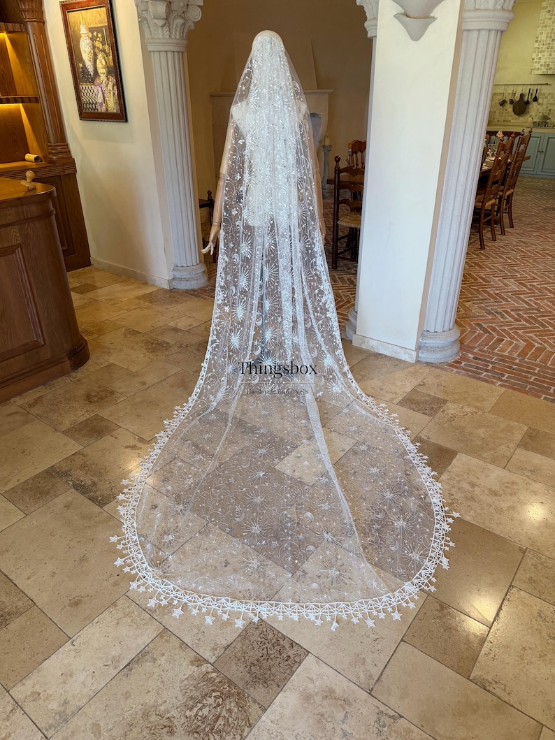 May include: A long, white, sheer bridal veil featuring intricate embroidery of stars, moons, and celestial patterns. The edges are finished with a delicate lace trim shaped like small stars. The text Thingsbox is printed on the fabric.