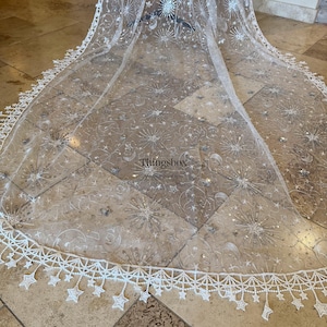 May include: A sheer white floor-length veil featuring silver embroidered celestial motifs including stars, moons, and sunbursts. The scalloped edge is adorned with dangling star-shaped lace appliqu&eacute;s. The text Thingsbox is visible in the center.