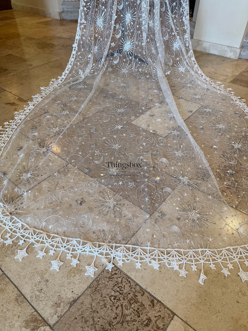 May include: A sheer white floor-length veil featuring silver embroidered celestial motifs including stars, moons, and sunbursts. The scalloped edge is adorned with dangling star-shaped lace appliqu&eacute;s. The text Thingsbox is visible in the center.