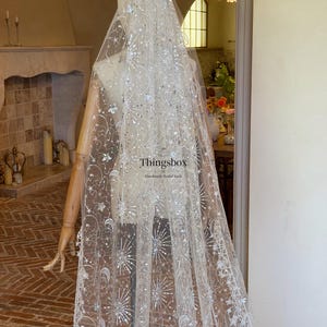 May include: A long, sheer white bridal veil featuring intricate silver embroidery of stars, crescent moons, and firework-like bursts. The fabric is embellished with shimmering sequins. The text "Thingsbox Handmade Bridal Veils" is visible.