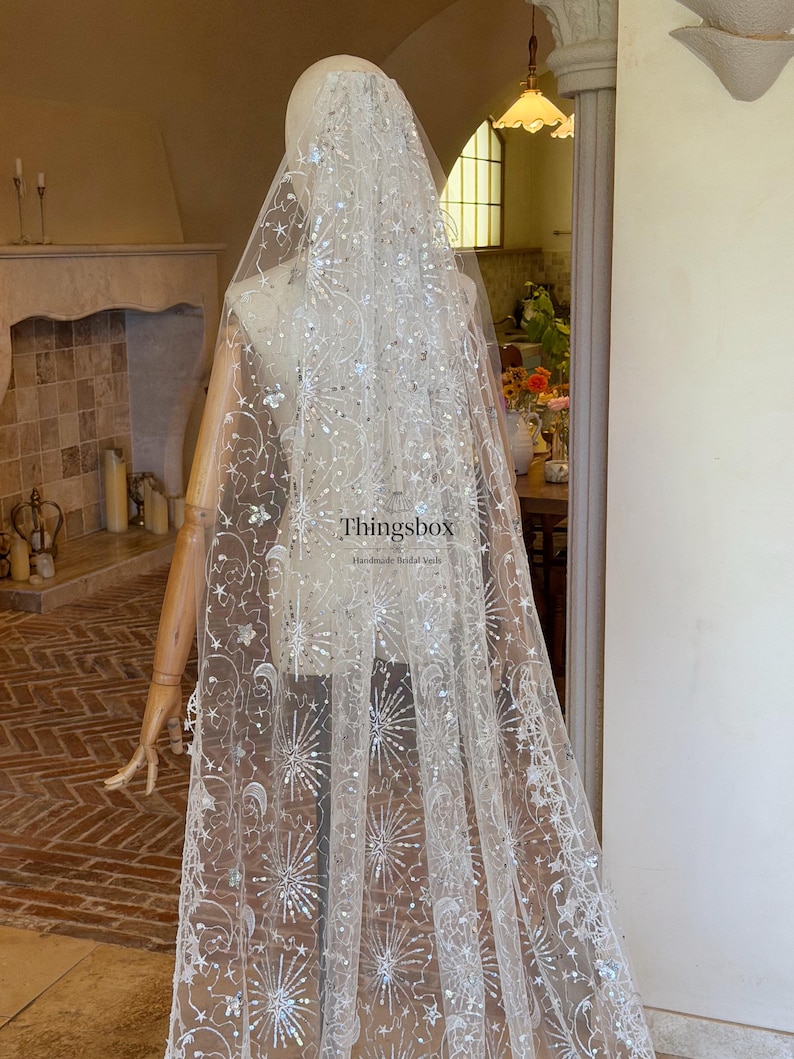 May include: A long, sheer white bridal veil featuring intricate silver embroidery of stars, crescent moons, and firework-like bursts. The fabric is embellished with shimmering sequins. The text "Thingsbox Handmade Bridal Veils" is visible.