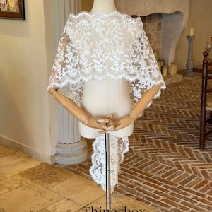 May include: A white lace bridal cape with a scalloped edge is displayed on a mannequin. The cape features an intricate floral pattern and a long, flowing train. The text "Thingsbox Handmade Bridal Veils" is visible at the bottom of the image.