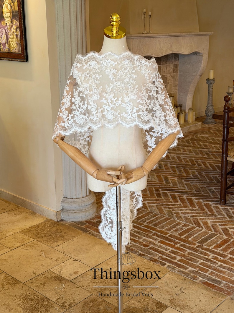 May include: A white lace bridal cape with a scalloped edge is displayed on a mannequin. The cape features an intricate floral pattern and a long, flowing train. The text "Thingsbox Handmade Bridal Veils" is visible at the bottom of the image.