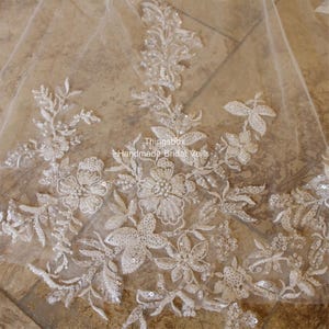 May include: Close-up of a white bridal veil with delicate floral lace and pearl embellishments. The lace features a floral pattern with small pearls and sequins. The text "Thingsbox - Handmade Bridal Veils" is visible in the lower right corner.