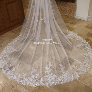 May include: A white bridal veil with a delicate lace trim. The veil is made of soft tulle and has a cathedral length. The lace trim features floral designs and is very intricate. The veil is displayed on a tiled floor. The text "Thingsbox -Handmade Bridal Veils- " is visible in the image.