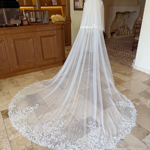 Puede incluir: Un velo de novia largo y blanco con un delicado ribete de encaje floral en el borde inferior. El velo está hecho de una tela transparente y fluida y está diseñado para ser usado durante una ceremonia de boda. El logotipo de Thingsbox es visible en la parte inferior.