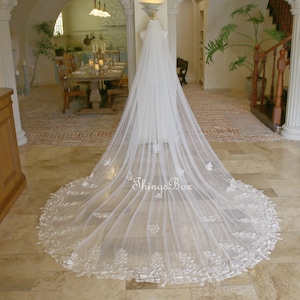 Puede incluir: Un velo de novia blanco largo con delicado ribete de encaje floral. El velo se exhibe en un maniquí, mostrando su longitud y diseño intrincado. El texto "Things Box" es visible en el velo.