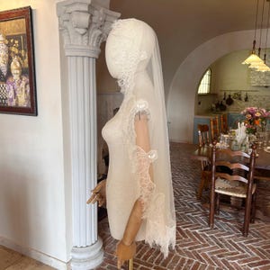 May include: A long, ivory-colored bridal veil draped over a mannequin head and shoulders. The veil is made of sheer tulle fabric and features intricate, scalloped floral lace trim along the edges that extends down the length of the veil.