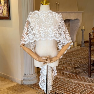 May include: A white lace bridal cape with floral patterns is displayed on a mannequin. The cape features a scalloped edge and a long, flowing train. The mannequin has wooden arms and a gold-colored head. The text "Thingsbox Handmade Bridal Veils" is visible.