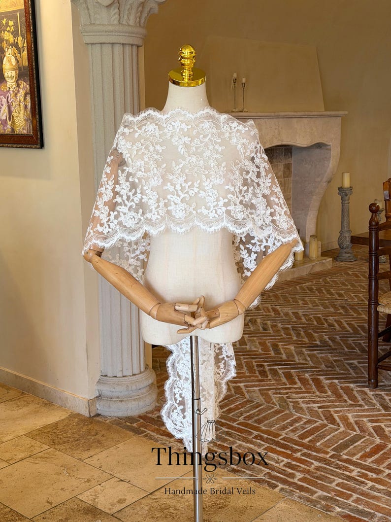 May include: A white lace bridal cape with floral patterns is displayed on a mannequin. The cape features a scalloped edge and a long, flowing train. The mannequin has wooden arms and a gold-colored head. The text "Thingsbox Handmade Bridal Veils" is visible.