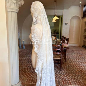 Może przedstawiać: Długi, kremowy welon ślubny z delikatnej koronki, narzucony na manekin. Welon ma skomplikowane kwiatowe wzory na brzegach. Widoczny jest tekst "Thingsbox - Handmade Bridal Veils". Scena jest w jasnym pokoju.
