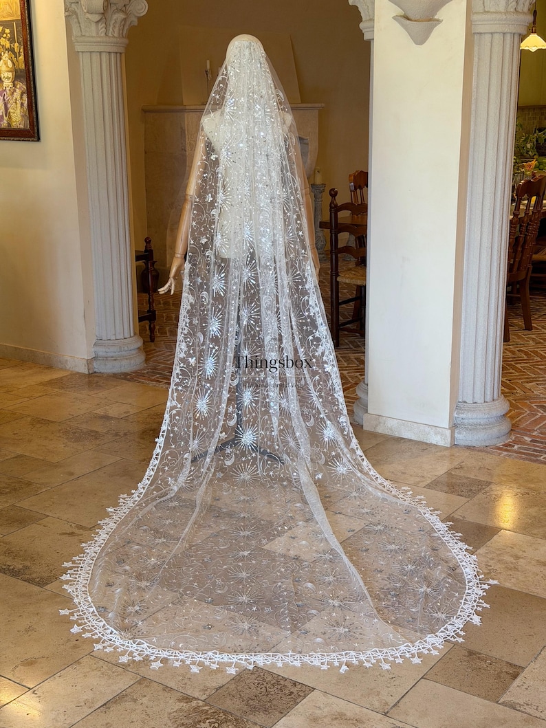 May include: A long, floor-length white bridal veil made of sheer tulle, featuring intricate silver embroidery of stars, moons, and sunburst patterns. The edges are finished with a delicate lace trim of small star shapes. The text Thingsbox is visible.