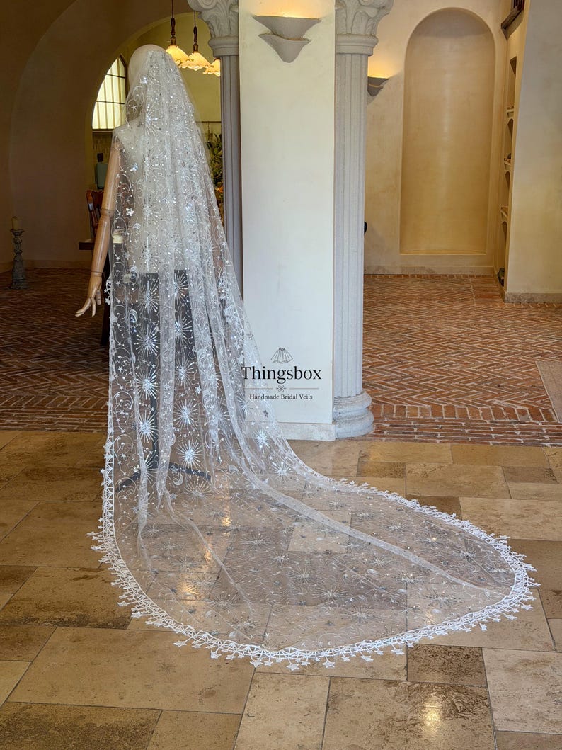 May include: A long, sheer white bridal veil featuring intricate silver embroidery of stars, moons, and celestial patterns. The veil has a scalloped edge with small star-shaped lace details and is displayed on a mannequin. Text reads Thingsbox.