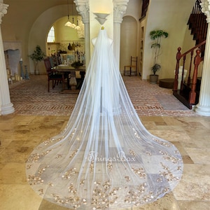 Pode incluir: Um véu de noiva de tule branco com detalhes florais dourados. O véu é longo e fluido, com uma borda festoneada. O véu é apresentado em um manequim em uma sala com piso de ladrilho.