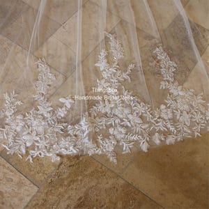 May include: A white bridal veil with delicate floral lace trim. The lace features small white beads and sequins. The veil is made of soft tulle and has a sheer, flowing look. The text "Thingsbox - Handmade Bridal Veils" is visible in the image.