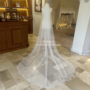 Könnte beinhalten: Ein langer, durchsichtiger weißer Brautschleier mit gewelltem Rand, verziert mit kleinen, zarten Perlen. Der Schleier wird auf einer Schaufensterpuppe präsentiert, mit dem Text "Thingsbox - Handmade Bridal Veils" sichtbar.