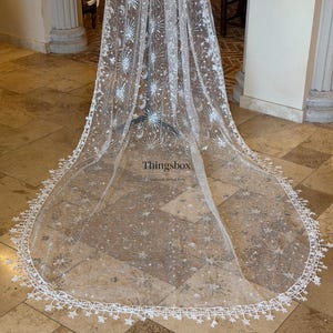 May include: A long, sheer white bridal veil featuring an intricate silver embroidered pattern of stars, moons, and sunbursts. The edges are finished with a delicate white lace trim shaped like small stars. The text Thingsbox is visible.