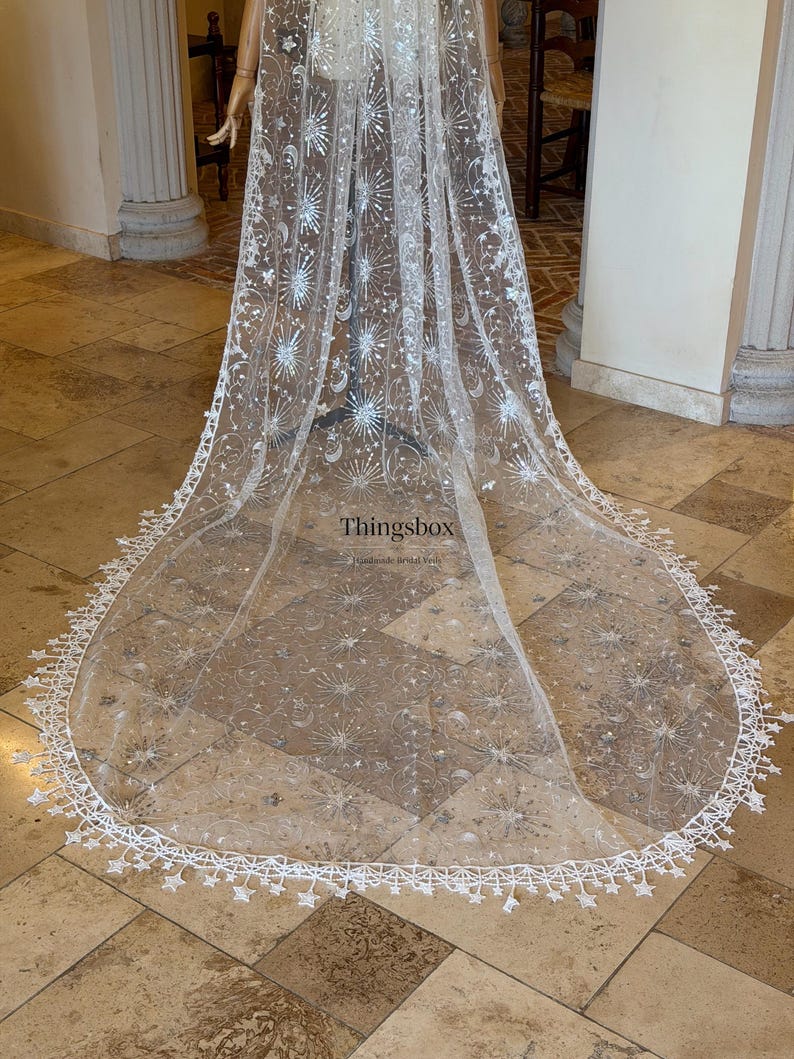 May include: A long, sheer white bridal veil featuring an intricate silver embroidered pattern of stars, moons, and sunbursts. The edges are finished with a delicate white lace trim shaped like small stars. The text Thingsbox is visible.