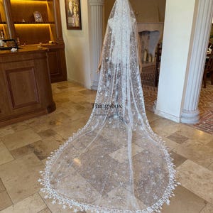 May include: A long, floor-length white bridal veil featuring intricate embroidery of celestial motifs including stars and sunbursts, accented with sequins and a scalloped lace edge with star-shaped details. The text Thingsbox is visible.