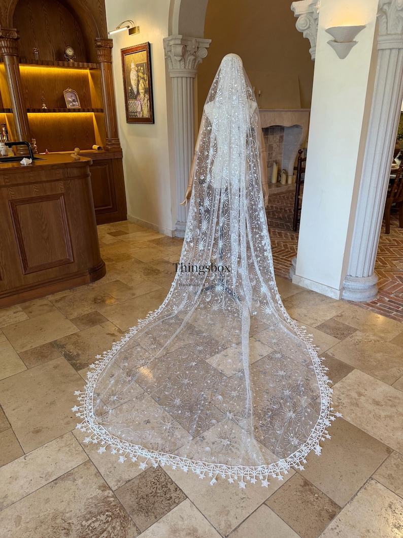 May include: A long, floor-length white bridal veil featuring intricate embroidery of celestial motifs including stars and sunbursts, accented with sequins and a scalloped lace edge with star-shaped details. The text Thingsbox is visible.