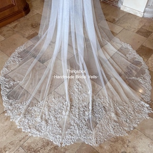 May include: A long, sheer white bridal veil with intricate floral lace detailing along the bottom edge. The veil is made of a delicate, flowing fabric and features the text "Thingsbox - Handmade Bridal Veils".