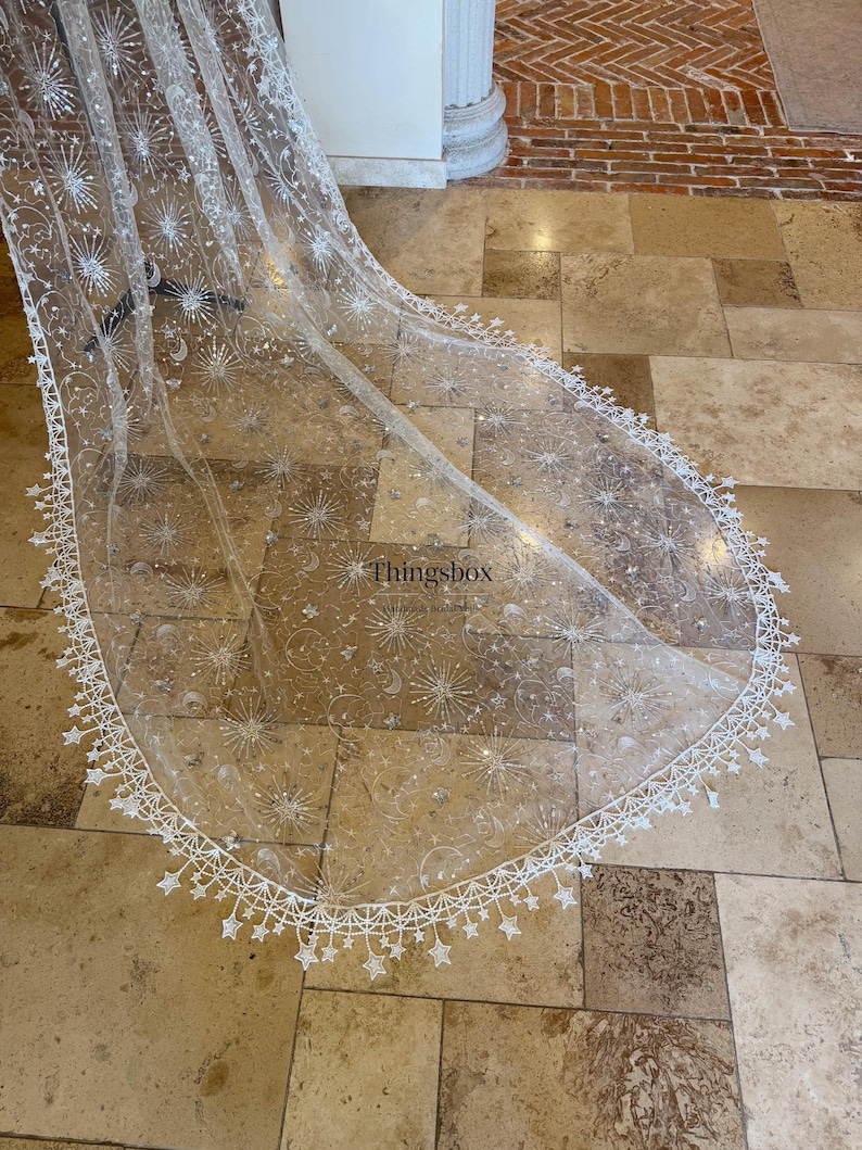 May include: A long, sheer white bridal veil featuring intricate silver embroidery of celestial motifs including stars, moons, and sunbursts. The edges are finished with a delicate white lace trim shaped like small stars. The text Thingsbox is visible.
