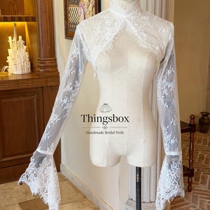 Op de afbeelding: Een witte kanten bruidsbolero met lange mouwen en uitlopende manchetten wordt op een mannequin getoond. De bolero heeft een hoge halslijn en ingewikkelde bloemenkantpatronen. De tekst "Thingsbox Handmade Bridal Veils" is zichtbaar.