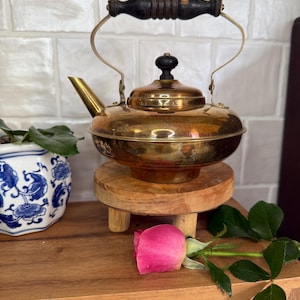 May include: A vintage brass teapot with a dark wooden handle and lid knob. The teapot sits on a small wooden stand. A pink rose and a blue and white patterned pot with a plant are also in the image.