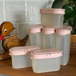 May include: A collection of clear plastic spice containers with light pink lids. The containers are rectangular and have a shaker top. A wooden duck figurine is visible on a wooden shelf in the background. The containers are designed for kitchen use.