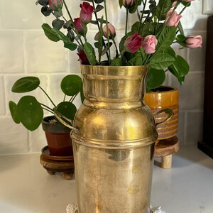 May include: A polished brass milk can vase with a cylindrical body, a flared neck, and two small side handles. The metal surface features a warm, metallic gold tone with subtle variations in texture and a smooth, reflective finish.