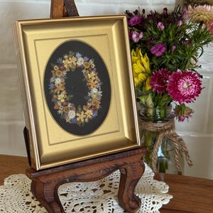May include: A framed floral artwork with a gold frame, displayed on a small wooden easel. The artwork features a wreath of dried flowers on a black background. A bouquet of colorful flowers sits in a glass jar nearby.