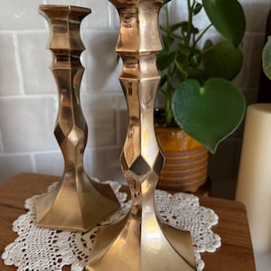 May include: Two gold-colored metal candlesticks with geometric designs. The candlesticks have a square base and a flared top, and are placed on a wooden surface with a white lace doily.