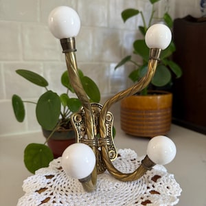 May include: A wall-mounted coat hook featuring a brass base with an intricate textured pattern and three curved brass arms. Each arm terminates in a smooth, round white ceramic knob. The metal has a warm, aged patina finish throughout the structure.