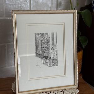 May include: Framed black and white print of a street scene. The artwork is in a gold-colored frame with a white mat. The print depicts a building with a brick facade and outdoor seating.