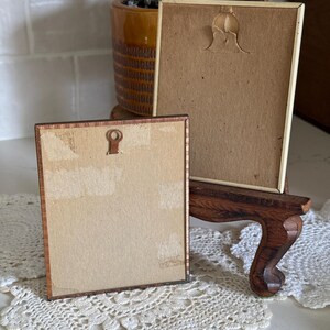 May include: Two small, vintage picture frames with brown wooden frames and tan backing. One frame is resting on a small wooden easel. The frames are displayed on a white lace doily. A potted plant is in the background.