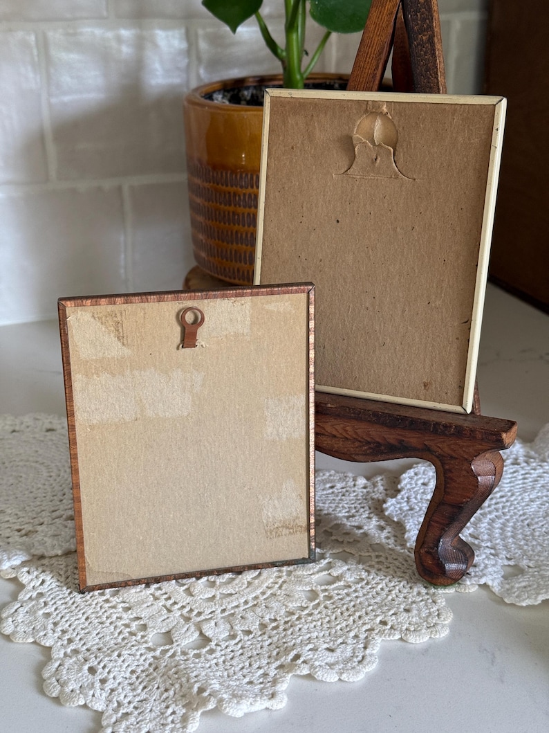 May include: Two small, vintage picture frames with brown wooden frames and tan backing. One frame is resting on a small wooden easel. The frames are displayed on a white lace doily. A potted plant is in the background.