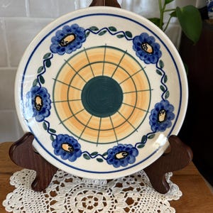 May include: A decorative ceramic plate with a floral design. The plate features a white background with blue flowers, green leaves, and a yellow and green center. The plate is displayed on a wooden stand.