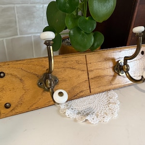 May include: A rectangular oak wood wall rack featuring two brass metal hooks. Each hook is equipped with two smooth white ceramic knobs at the ends. The wooden base has a visible grain pattern and includes pre-drilled holes for mounting purposes.