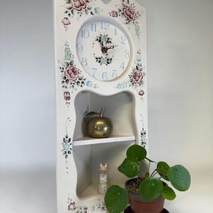 May include: White corner clock with a floral design. The clock face has blue numerals and black hands. A shelf holds a brass apple and a figurine. A potted plant sits nearby. The clock is approximately 24 inches tall.