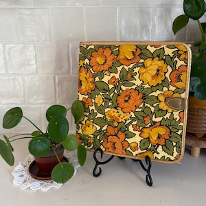 May include: A square zippered portfolio featuring a vibrant floral pattern with orange, yellow, and brown flowers against a cream background. The case has a gold-toned metallic trim and a small tab closure on the right side, displayed on a black stand.