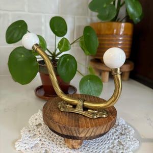 May include: A double-pronged brass wall hook featuring two white spherical finials at the ends of the curved arms. The hook is mounted onto a circular, dark-stained wooden base with a visible grain pattern and a small central support leg underneath.
