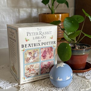 May include: A boxed set of "The Peter Rabbit Library by Beatrix Potter" books. The box is white with illustrations and text. A light blue, round trinket box with white rabbit designs sits on a white lace doily. Green plants are in the background.