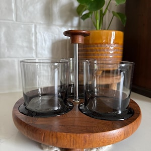 May include: A vintage bar set featuring a wooden lazy susan with two gray-tinted glasses. The set includes a central metal and wood handle. The glasses sit on black bases within the wooden tray. A decorative piece for serving drinks.