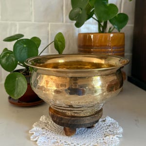 May include: A polished brass bowl with a hammered texture and a wide, rounded rim. The bowl is supported by a circular, dark brown wooden base featuring three small, sturdy feet, resting upon a white, circular crocheted lace doily.