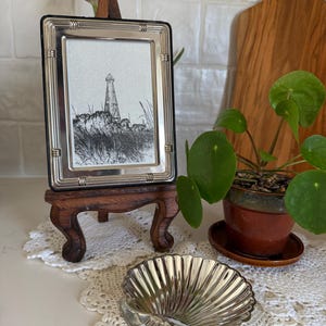 May include: A silver-framed picture of a lighthouse on a wooden stand, a silver seashell-shaped dish, and a potted plant with green leaves. The picture frame has a black border and is set on a small, ornate wooden easel. The seashell dish is on a white lace doily.