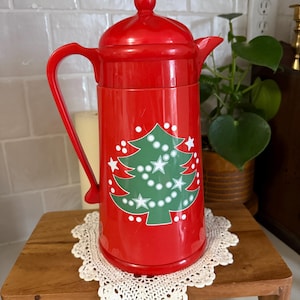 May include: A red vintage thermal carafe with a Christmas tree design. The carafe has a handle, a lid, and a spout. It is sitting on a wooden stand with a white lace doily.